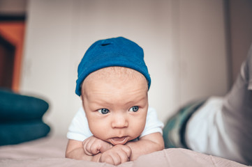 Cute little baby boy lying on pink blanket at bedroom. Newborn. Baby stays awake on the bed. Closeup portrait of emotional newborn baby in blue jeans, blue cap, and white t-shirt