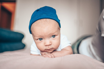 Cute little baby boy lying on pink blanket at bedroom. Newborn. Baby stays awake on the bed. Closeup portrait of emotional newborn baby in blue jeans, blue cap, and white t-shirt