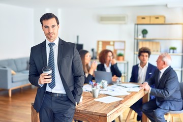 Group of business workers smiling happy and confident working together in a meeting. One of them, standing with smile on face looking at camera drinking coffee at the office.