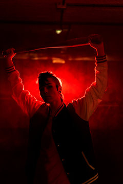 Rebel woman in casual jacket with piercing and modern hairstyle holding bat among colorful red light and steam