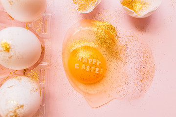 Top view of broken glittered egg with text Happy Easter and raw eggs in plastic box on pink background