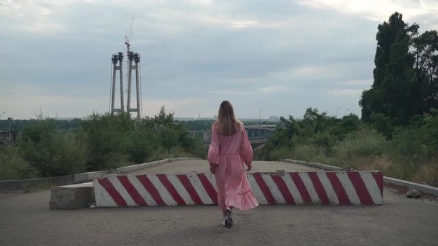 beautiful, young girl with long hair pink dress walks alone along asphalt road, past concrete red blocks, turns sharply. Against background unfinished bridges, lampposts, metal structures, cars, city
