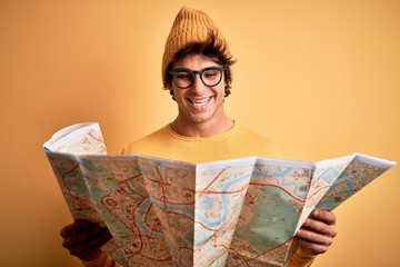 Young tourist man holding map wearing glasses and cap over isolated yellow background with a happy face standing and smiling with a confident smile showing teeth