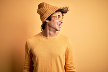 Young handsome man wearing casual t-shirt and glasses over isolated yellow background looking away to side with smile on face, natural expression. Laughing confident.