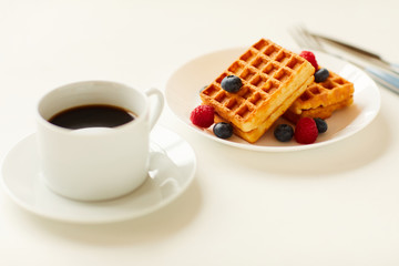 Obraz na płótnie Canvas Warm-toned close up of sweet dessert waffles with berry topping next to cup of black coffee on white background, copy space