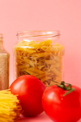 Noodles, rice, pasta in glass jars stand on a pink background. Nearby are red tomatoes and green cucumber. Raw materials for cooking.
