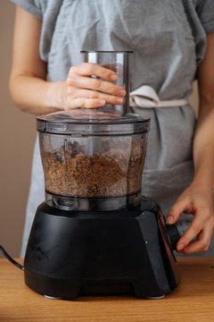Crop housewife in grey apron preparing food at wooden table and using food processor for grinding beans 