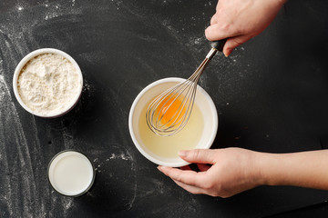 Female hands beat a broken egg to make dough on a table background with ingredients for homemade baking.