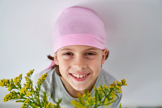 Cheerful little boy with cancer diagnosis wearing pink bandana and looking at camera while holding vase with flowers and standing at wall