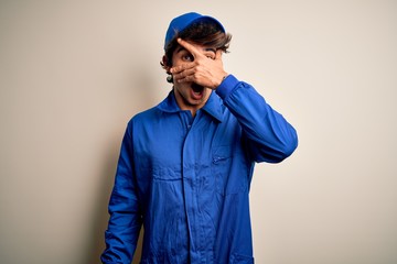 Young mechanic man wearing blue cap and uniform standing over isolated white background peeking in shock covering face and eyes with hand, looking through fingers with embarrassed expression.