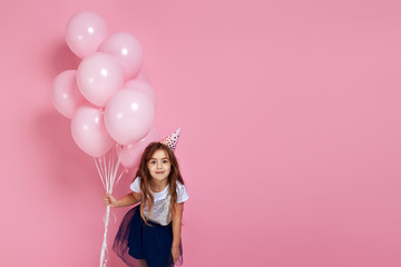 Beautiful happy little child girl in dress and birthday hat posing with pastel pink air balloons isolated on pink background. birthday party. copy space