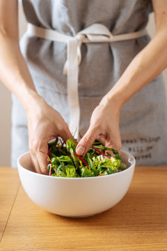 Crop female in apron mixing fresh herbs in bowl while cooking healthy vegan salad on table in kitchen