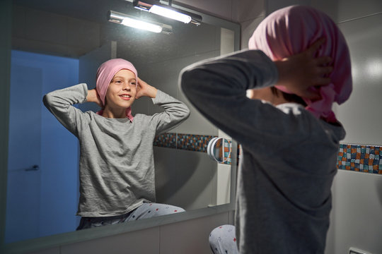 Back view of sick little child putting on pink bandana in front of mirror in bathroom