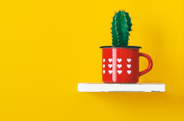 Shelf with little cactus plant potted in vintage red cup with hearts shape on yellow wall...