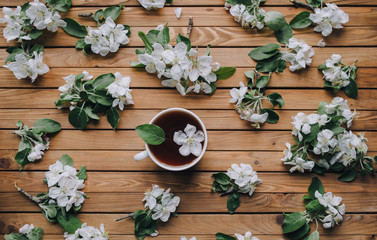 White cup of tea with petals of an apple tree on a brown wooden background, view from above.