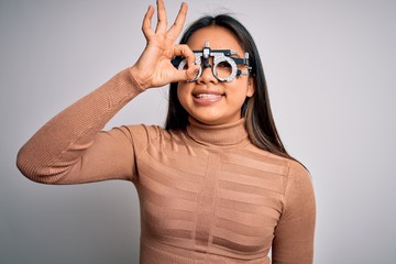 Young asian optical girl controlling eyesight using optometry glasses over white background doing ok gesture with hand smiling, eye looking through fingers with happy face.