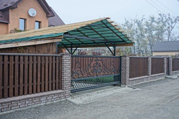 brown metal gate with a black forged pattern and a long fence of wooden boards and bricks on a rural street