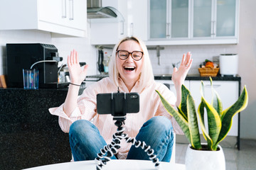 Happy girl sitting at home kitchen and holding videocall. Young woman using smartphone for video call with friend or family. Vlogger recording webinar. Woman looking camera and waving greeting hands