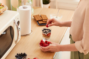 Warm-toned close up of unrecognizable young woman making yogurt parfait with fresh berries and...