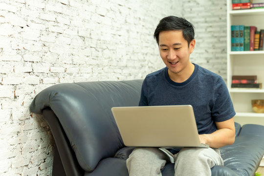 Young Smiling Asian Man In Casual Clothing On Sofa Working On Laptop Computer. Male Freelancer Working From Home On Notebook. Work From Home Concept