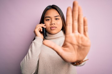 Young asian girl having conversation talking on the smartphone over isolated pink background with open hand doing stop sign with serious and confident expression, defense gesture