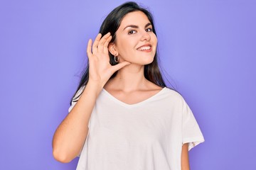 Young beautiful brunette woman wearing casual white t-shirt over purple background smiling with hand over ear listening an hearing to rumor or gossip. Deafness concept.