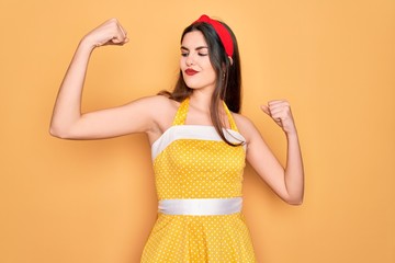 Young beautiful pin up woman wearing 50s fashion vintage dress over yellow background showing arms...