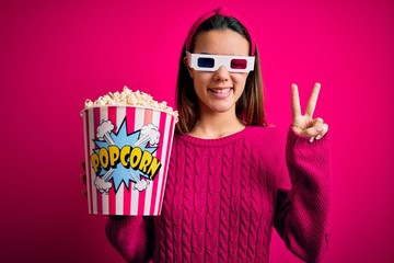 Obraz premium Young beautiful girl watching movie using 3d glasses eating box with popcorns smiling with happy face winking at the camera doing victory sign with fingers. Number two.