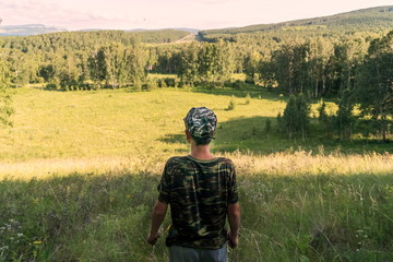 A boy in a shirt and cap, camouflage colors, stands in the shade and looks at the endless taiga on a summer sunny day.