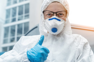 Portrait of tired exhausted female doctor, scientist or nurse wearing face mask and biological hazmat ppe suit showing comfidence thumbs up like gesture outdoor. Coronavirus covid-19 outbreak danger