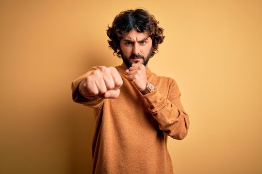 Young handsome man with beard wearing casual sweater standing over yellow background Punching fist to fight, aggressive and angry attack, threat and violence