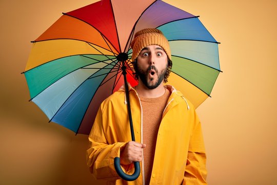 Young handsome man with beard wearing raincoat for rainy day holding colorful umbrella scared in shock with a surprise face, afraid and excited with fear expression