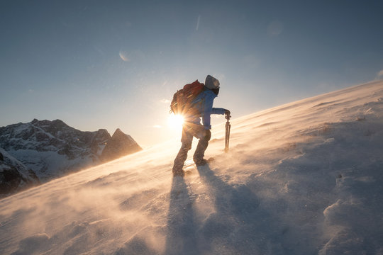 Man traveler with tripod are climbing on slope hill on Ryten mount in blizzard at sunset