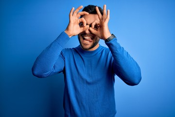 Naklejka premium Young handsome man with beard wearing casual sweater and glasses over blue background doing ok gesture like binoculars sticking tongue out, eyes looking through fingers. Crazy expression.