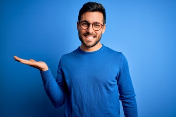 Young handsome man with beard wearing casual sweater and glasses over blue background smiling cheerful presenting and pointing with palm of hand looking at the camera.
