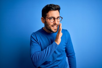 Young handsome man with beard wearing casual sweater and glasses over blue background hand on mouth telling secret rumor, whispering malicious talk conversation - obrazy, fototapety, plakaty