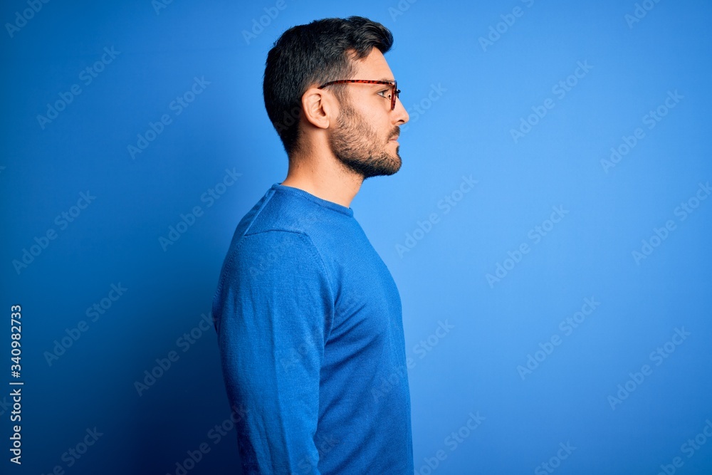 Poster young handsome man with beard wearing casual sweater and glasses over blue background looking to sid