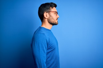 Young handsome man with beard wearing casual sweater and glasses over blue background looking to...