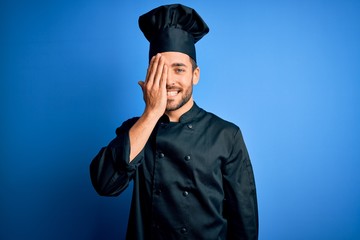 Young handsome chef man with beard wearing cooker uniform and hat over blue background covering one eye with hand, confident smile on face and surprise emotion.