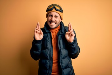 Young handsome skier man with beard wearing snow sportswear and ski goggles gesturing finger crossed smiling with hope and eyes closed. Luck and superstitious concept.
