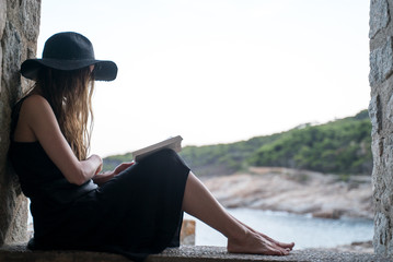 Girl reads book. A girl in a black hat and black silk dress sits in the niche of a fishing hut on the seashore and reads a book. Ocean view at a background. Studying outdoors, reading a novel.
