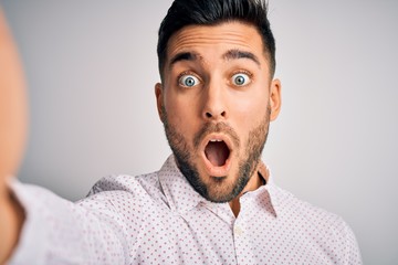 Young handsome man wearing shirt making selfie by the camera over white background scared in shock...