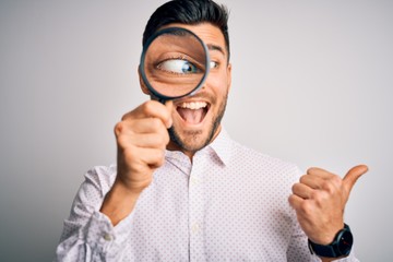 Fototapeta na wymiar Young detective man looking through magnifying glass over isolated background pointing and showing with thumb up to the side with happy face smiling