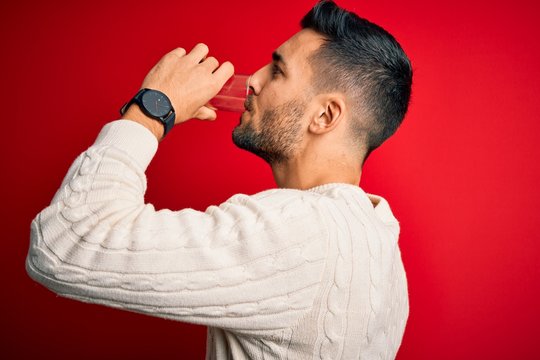 Young handsome man drinking glass of healthy water to refreshment standing over isolated red background