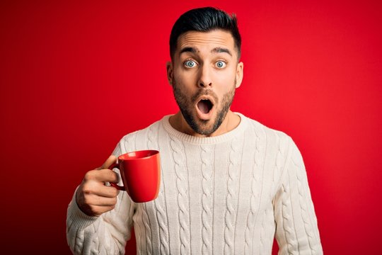 Young handsome man drinking a cup of hot coffee over red isolated background scared in shock with a surprise face, afraid and excited with fear expression