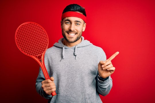 Young handsome sportsman holding tennis racket wearing sportswear over red background very happy pointing with hand and finger to the side