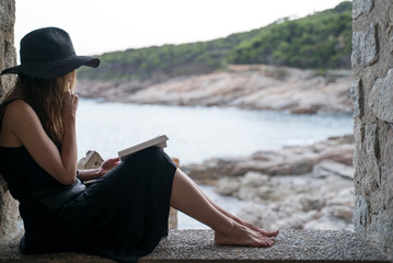 Girl reads book. A girl in a black hat and black silk dress sits in the niche of a fishing hut on the seashore and reads a book. Ocean view at a background. Studying outdoors, reading a novel.