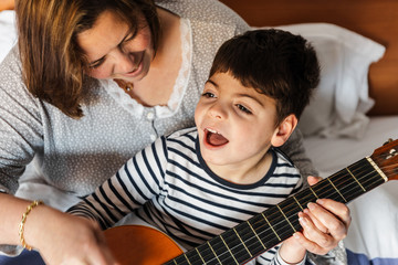 niño juega y aprende con  una mujer a tocar la guitarra en la cama. son madre e hijo, él tiene...