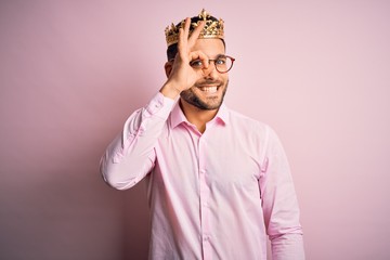 Young handsome business man wearing golden crown as king over pink background doing ok gesture with hand smiling, eye looking through fingers with happy face.