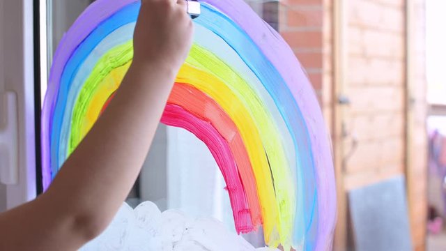 Rainbow drawing on the window. A little girl holds in her hands a large box with paints and draws. Good pictures and actions during the quarantine period. Hope for a brighter future. Soft focus